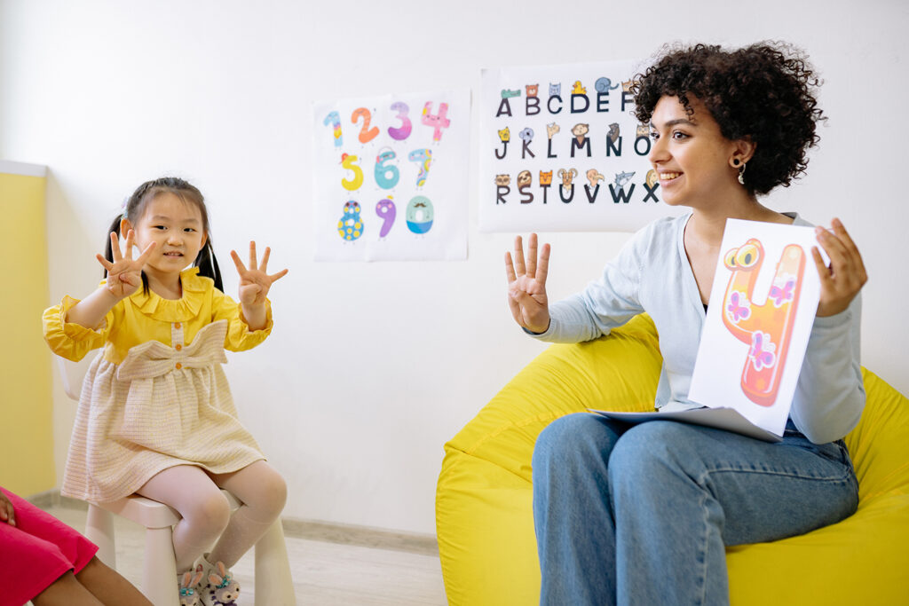 Woman preschool teacher sitting on a yellow beanbag chair and holding a sign that says 4. She also has four fingers held up to demonstrate for a child in her class who is also holding four fingers up to match.