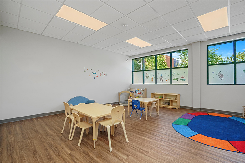 Therapeutic preschool or kindergarten classroom from our West Loop Chicago location. There are two child-sized tables with chairs, a brightly colored rug, a bookshelf filled with kids books, and a wall filled with windows that has stickers of animals on them.