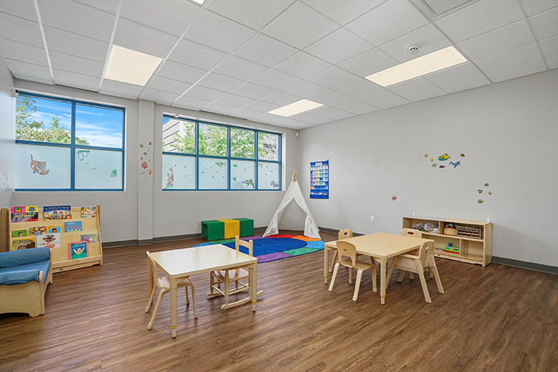 Therapeutic preschool or kindergarten classroom from our West Loop Chicago location. There are two child-sized tables with chairs, a brightly colored rug, a bookshelf filled with kids books, a child-sized couch, colorful block chairs, and a wall filled with windows that has stickers of animals on them.