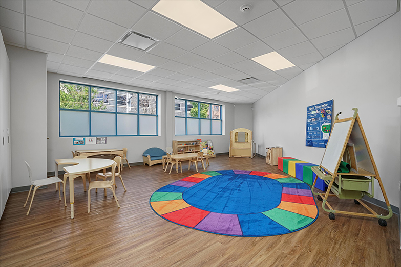 Therapeutic classroom with a big rainbow rug, tables and chairs for children, and a whiteboard. Therapeutic preschool or kindergarten classroom from our West Loop Chicago location. There are three child-sized tables with chairs, a brightly colored rug, a whiteboard with markers, child-sized cube seats in many colors, a child sized couch, a puppet station, and a wall filled with windows.
