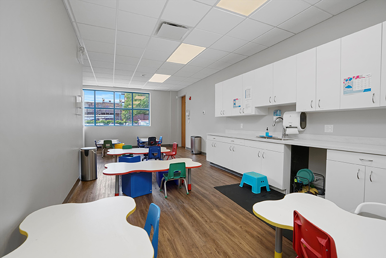 Therapeutic preschool and kindergarten lunchroom at our West Loop Chicago location. There are six child-sized tables with chairs throughout the room. On the right side are white cabinets.