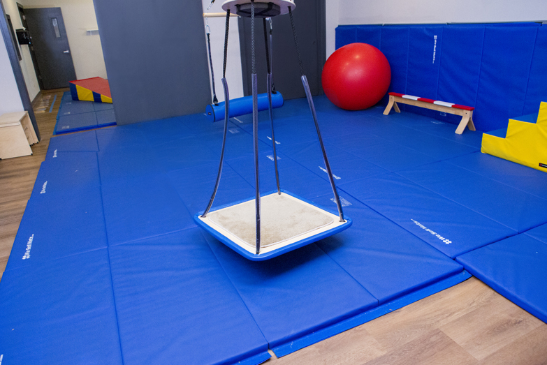 Interactive Therapeutic Sensorimotor gym at our Northcenter Chicago location. Contains a platform swing, an exercise ball, a balance beam, a bolster swing, walking steps, and a foam wedge. The floors are covered with blue padding for safety.