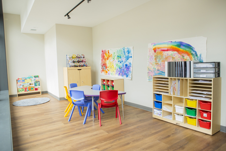 Therapeutic preschool or kindergarten classroom from our Northcenter Chicago location. There's a child-sized table, a bookshelf filled with kids books, a cabinet filled with colorful plastic tubs, and rainbow paintings on the walls.