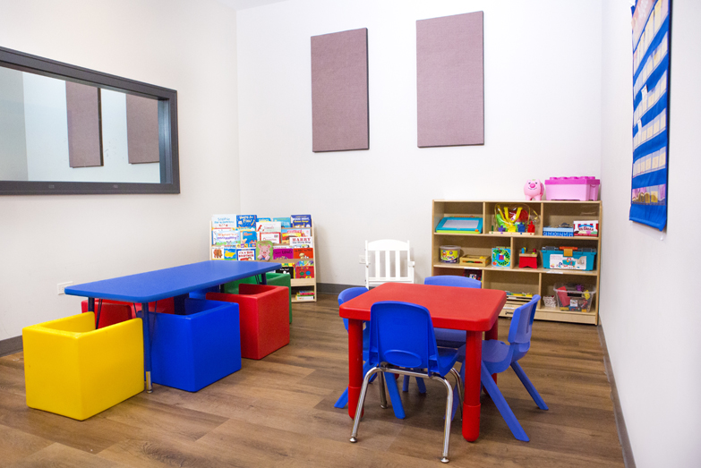 Therapeutic preschool or kindergarten classroom from our Northcenter Chicago location. There are two child-sized tables with brightly colored cube chairs, a bookshelf filled with kids books, a cabinet filled with toys, and a white rocking chair.