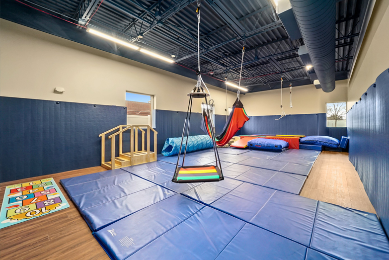 Interactive Therapeutic Sensorimotor gym at our Wheaton Illinois location. Includes a therapeutic rainbow platform swing for vestibular and proprioceptive input, a set of climbing steps with a handrail for balance, crash pads and mats for safety and deep pressure input surrounding a large rainbow colored trampoline, a long blue tunnel, and a hopscotch game. There is blue padding on the walls and floor for safety.