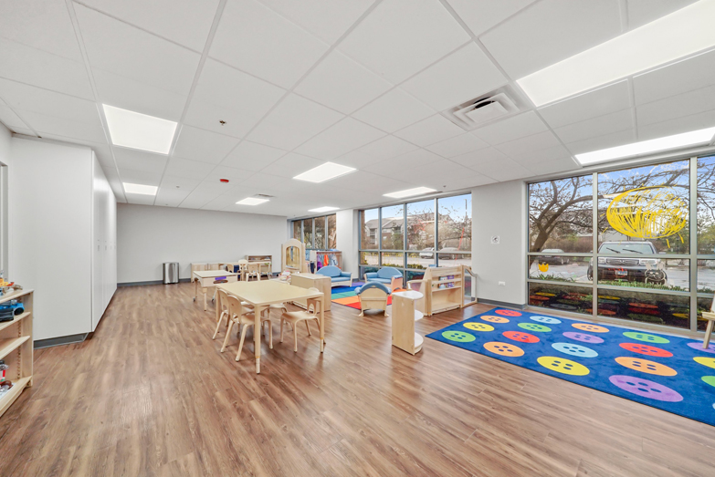 Therapeutic preschool or kindergarten classroom from our Wheaton Illinois location. There are several child-sized tables with chairs, a brightly colored rug, a bookshelf filled with kids books, and a wall full of windows looking outside.