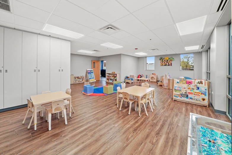 Therapeutic preschool or kindergarten classroom from our Wheaton Illinois location. There are three child-sized tables with chairs, a brightly colored rug with color cube chairs on it, a bookshelf filled with kids books, and a sensory table.