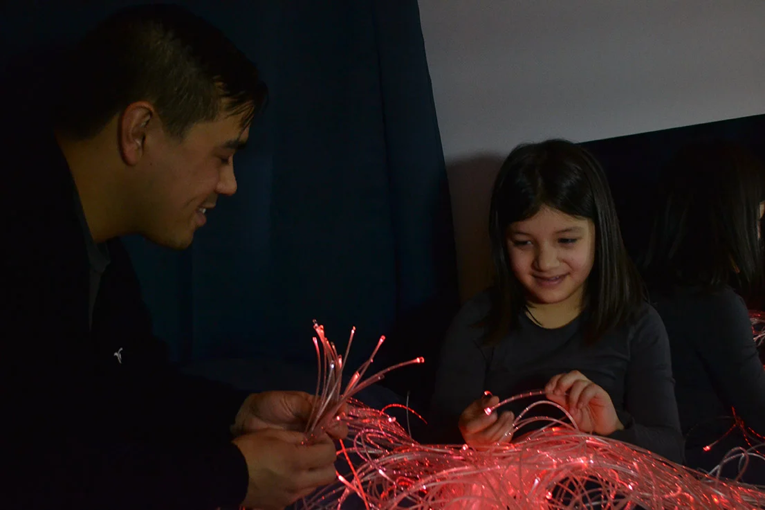 Child and man sitting in a dark sensory room playing with colorful lights